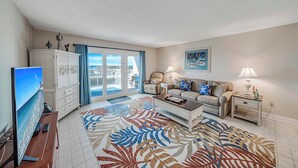 A spacious living room with a flat-screen TV, a white cabinet, a beige sofa, a patterned rug, and a view through the glass door.