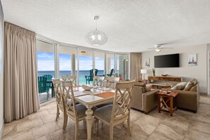 A spacious living area with a dining table, sofa, and TV. Large windows offer a view of the ocean.