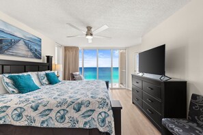 A bedroom with a bed, a TV, a ceiling fan, a window with a view of the ocean, and a painting on the wall.