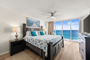 A bedroom with a bed, nightstands, a ceiling fan, a large window with a view of the ocean, and a painting on the wall.