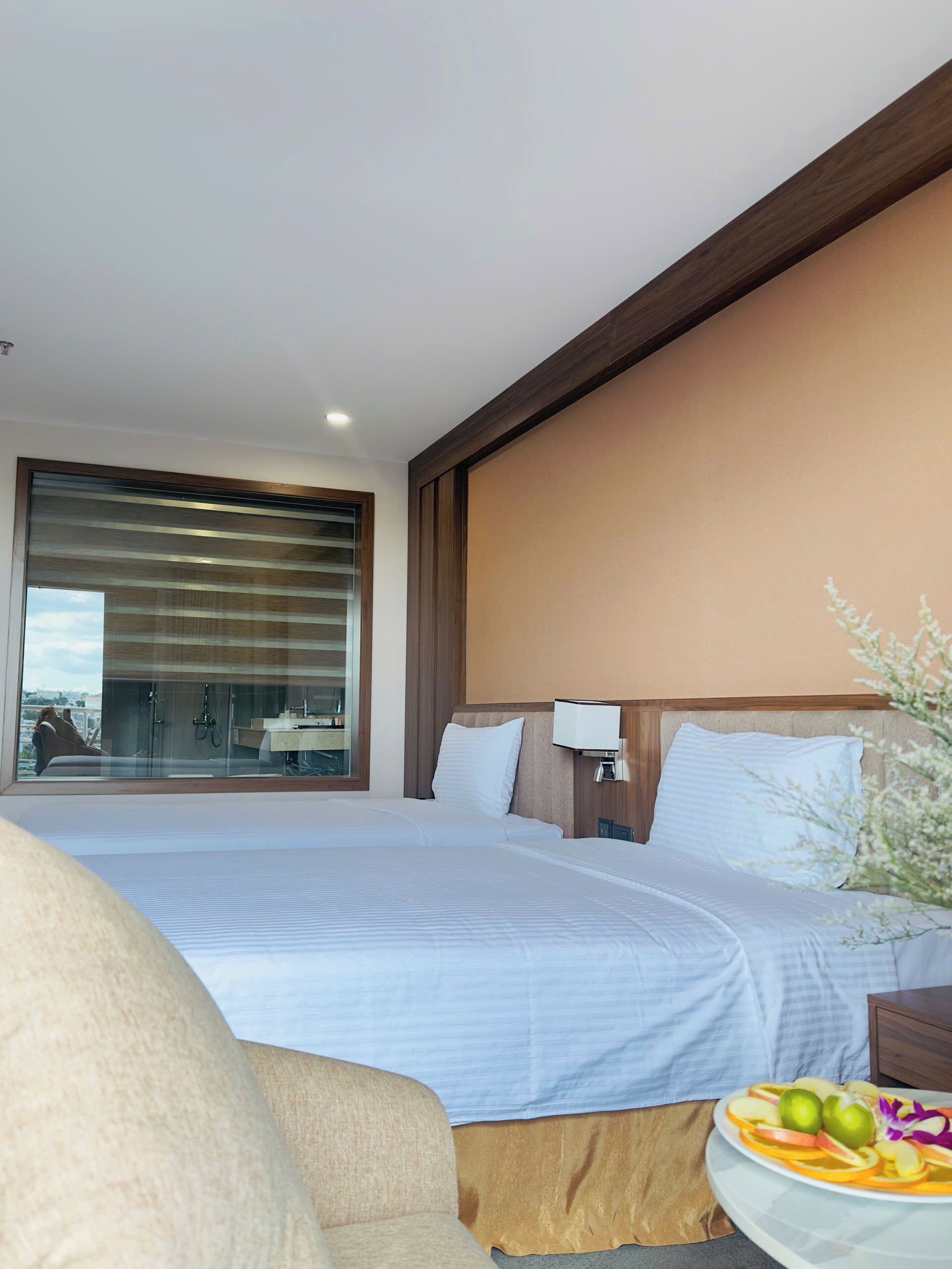 A modern hotel room with a large bed, a sofa, a small table with fruit, and a view of the city through a large window.