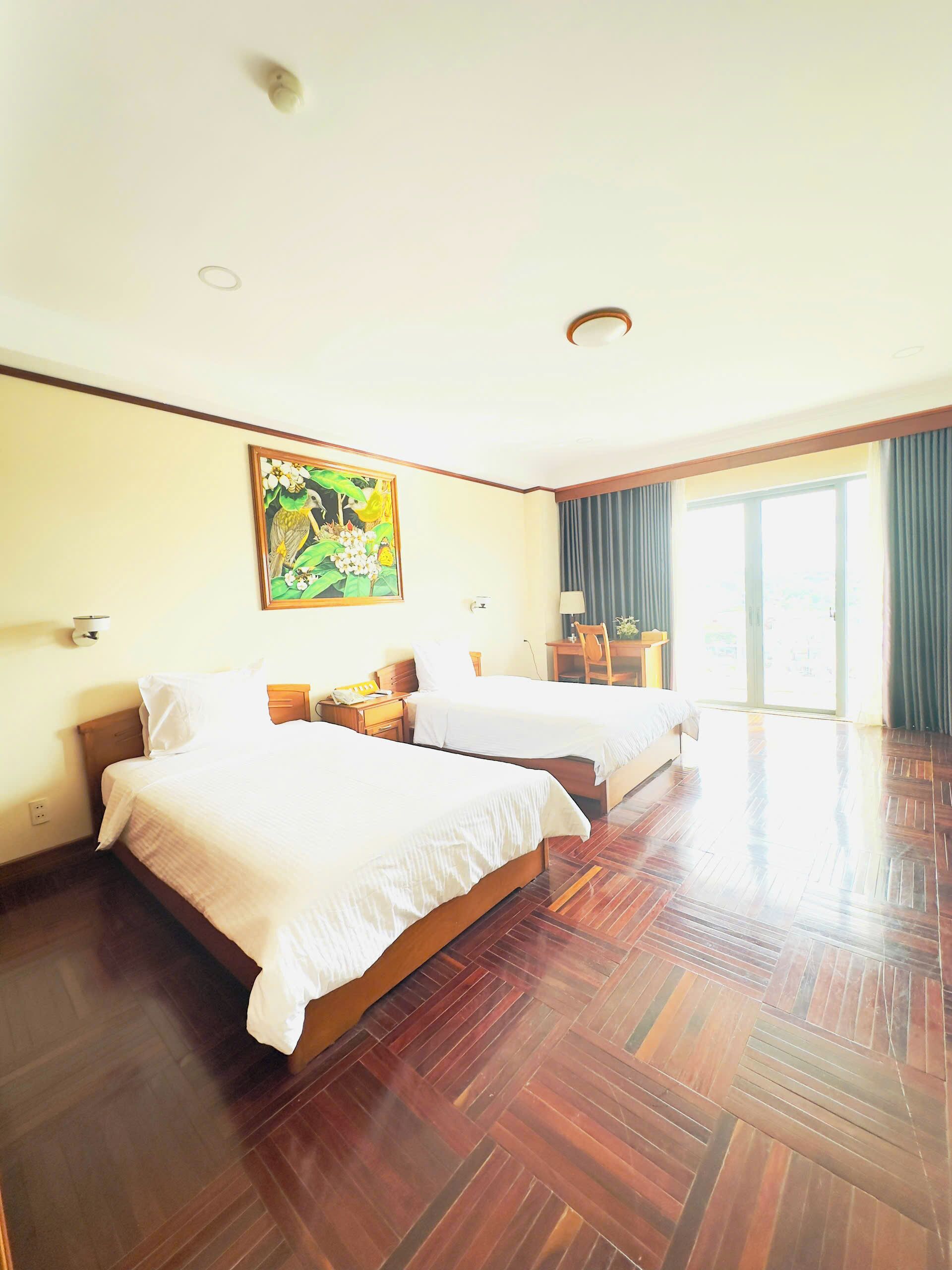 A hotel room with two beds, wooden flooring, a painting on the wall, and a large window with curtains.