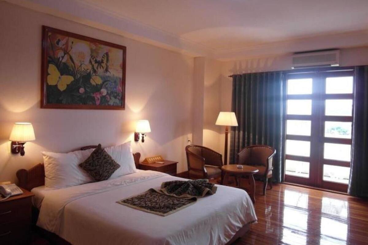 Une chambre d’hôtel avec un lit, deux lampes de chevet, un tableau, une chaise, une table et une fenêtre avec des rideaux.