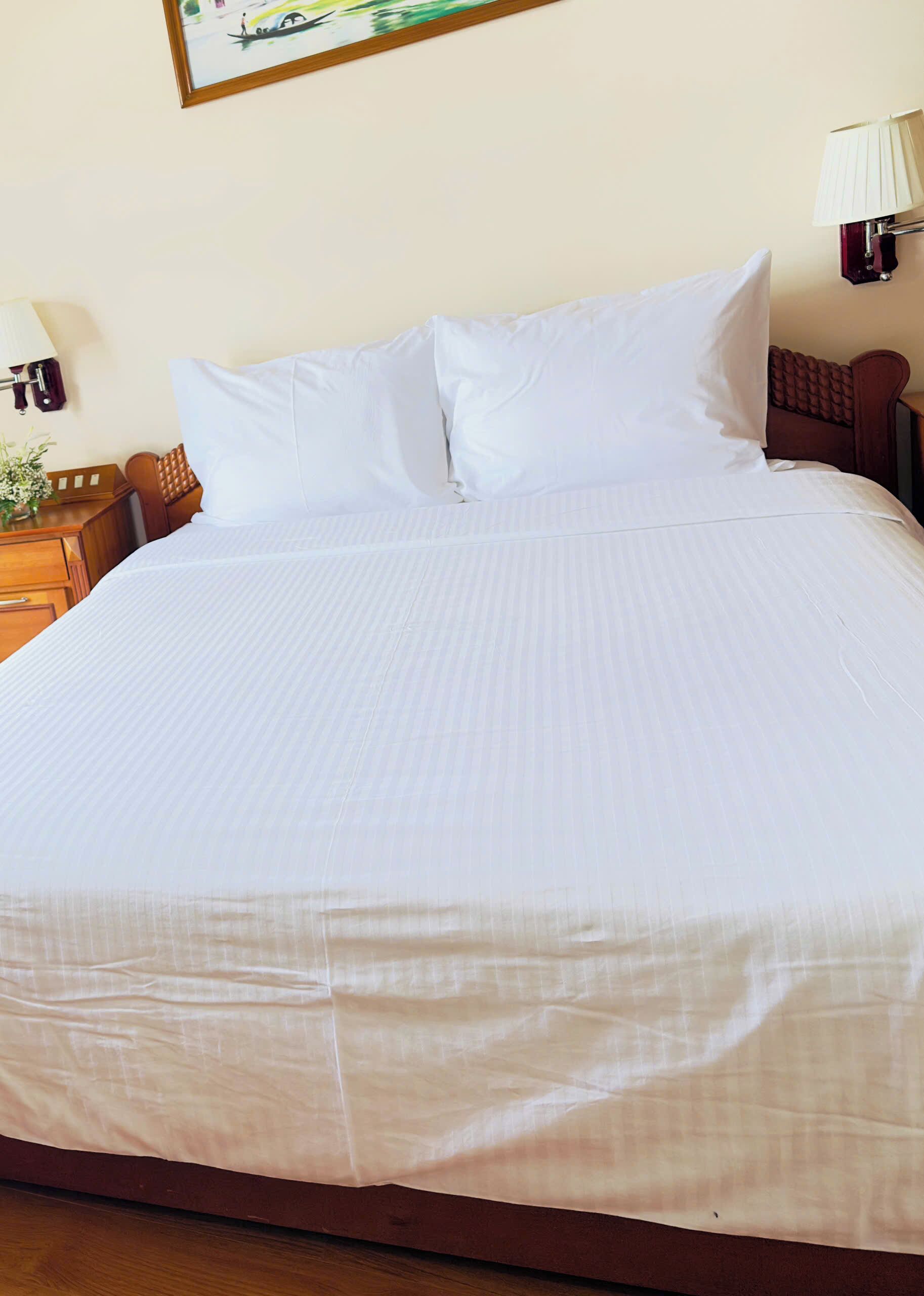 Una cama bien hecha con sábanas blancas, un cabecero de madera y una mesita de noche con una lámpara y un florero.