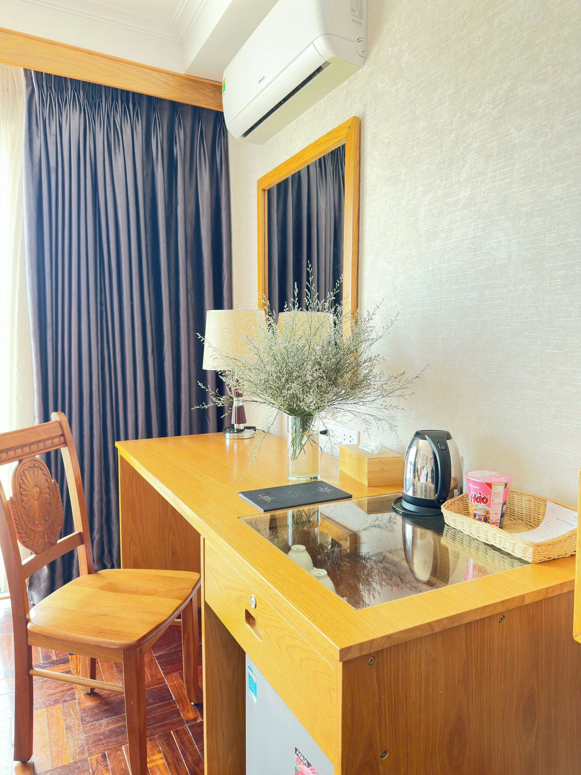 A hotel room with a wooden desk, a chair, a vase with dried flowers, a lamp, a kettle, and a basket with a container.