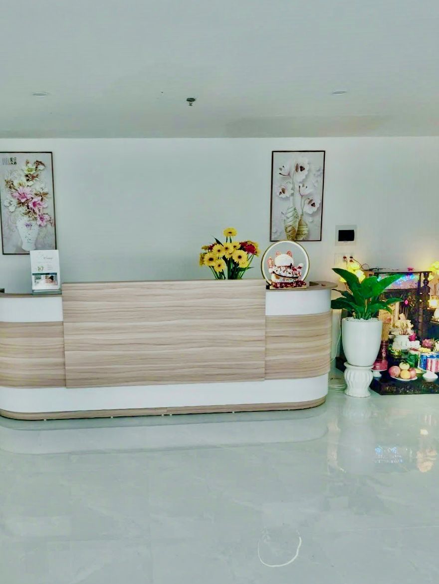 Un mostrador de recepción con acabado en madera clara, una obra de arte floral enmarcada encima y una planta en maceta a la derecha.