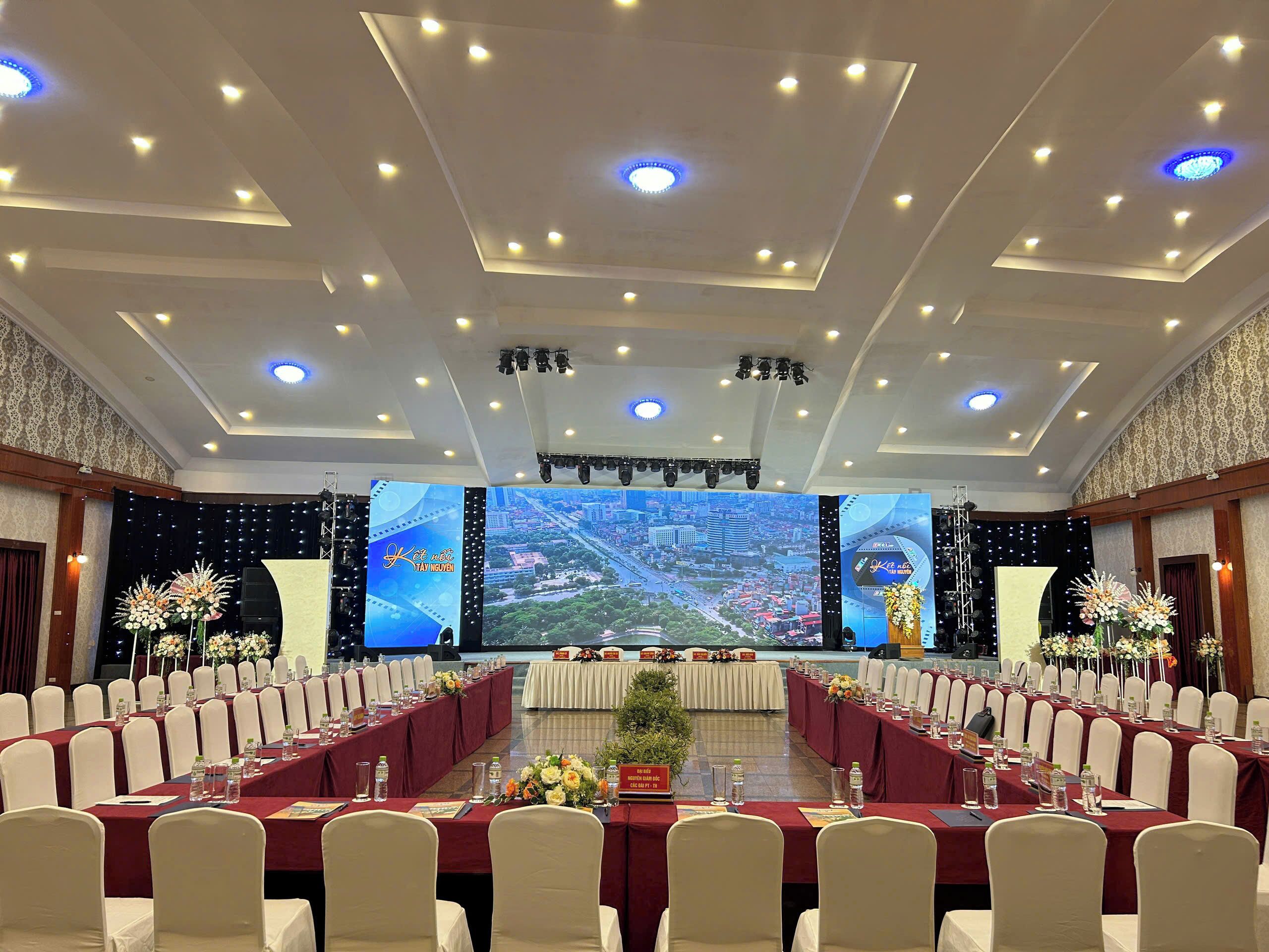 Une salle de conférence dotée d’un grand écran affichant une vue panoramique de la ville, de plusieurs tables avec des chaises et d’une scène équipée de matériel d’éclairage.