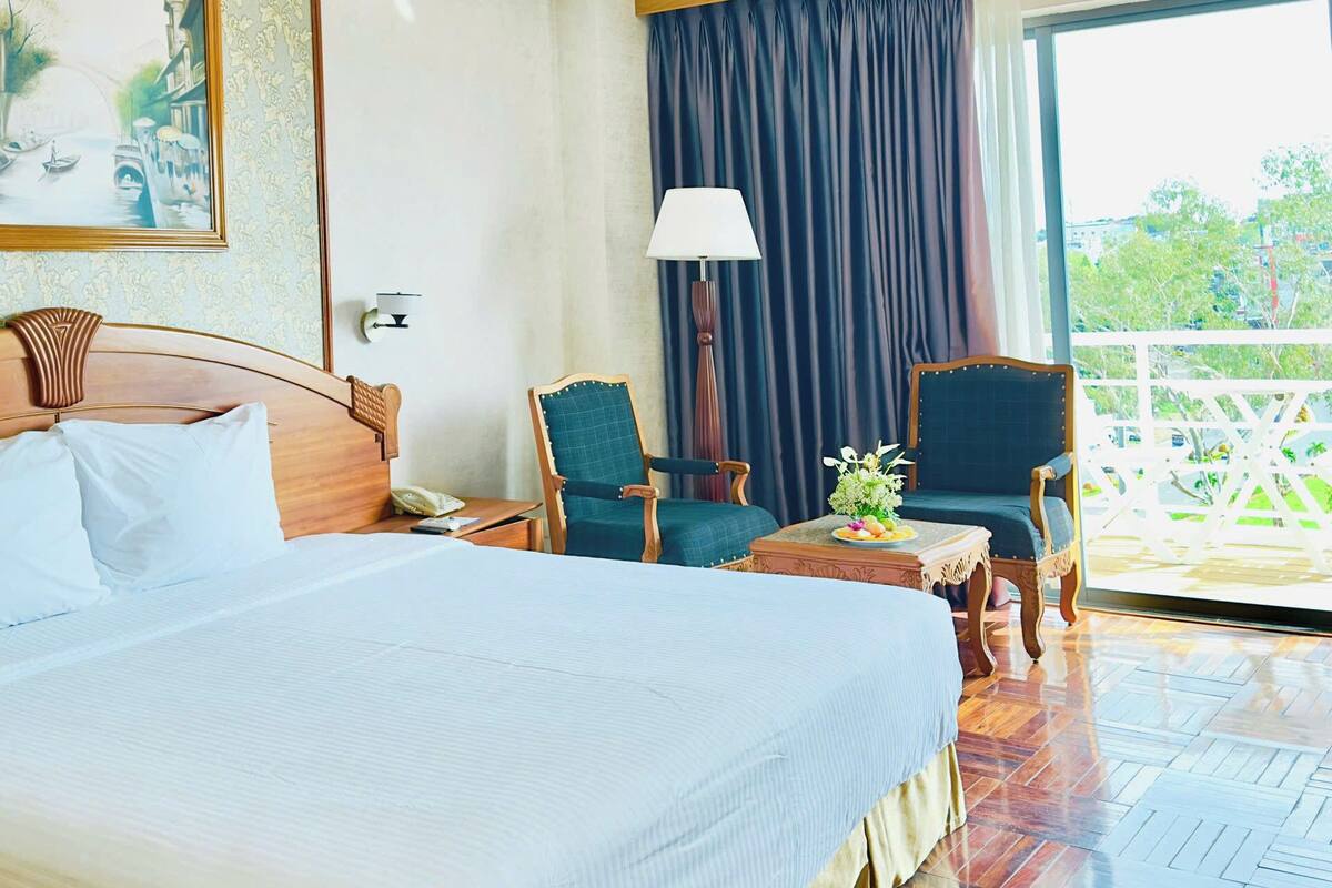 Une chambre d’hôtel avec un grand lit, deux fauteuils, une table avec un vase, une lampe et une vue sur la verdure depuis un balcon.