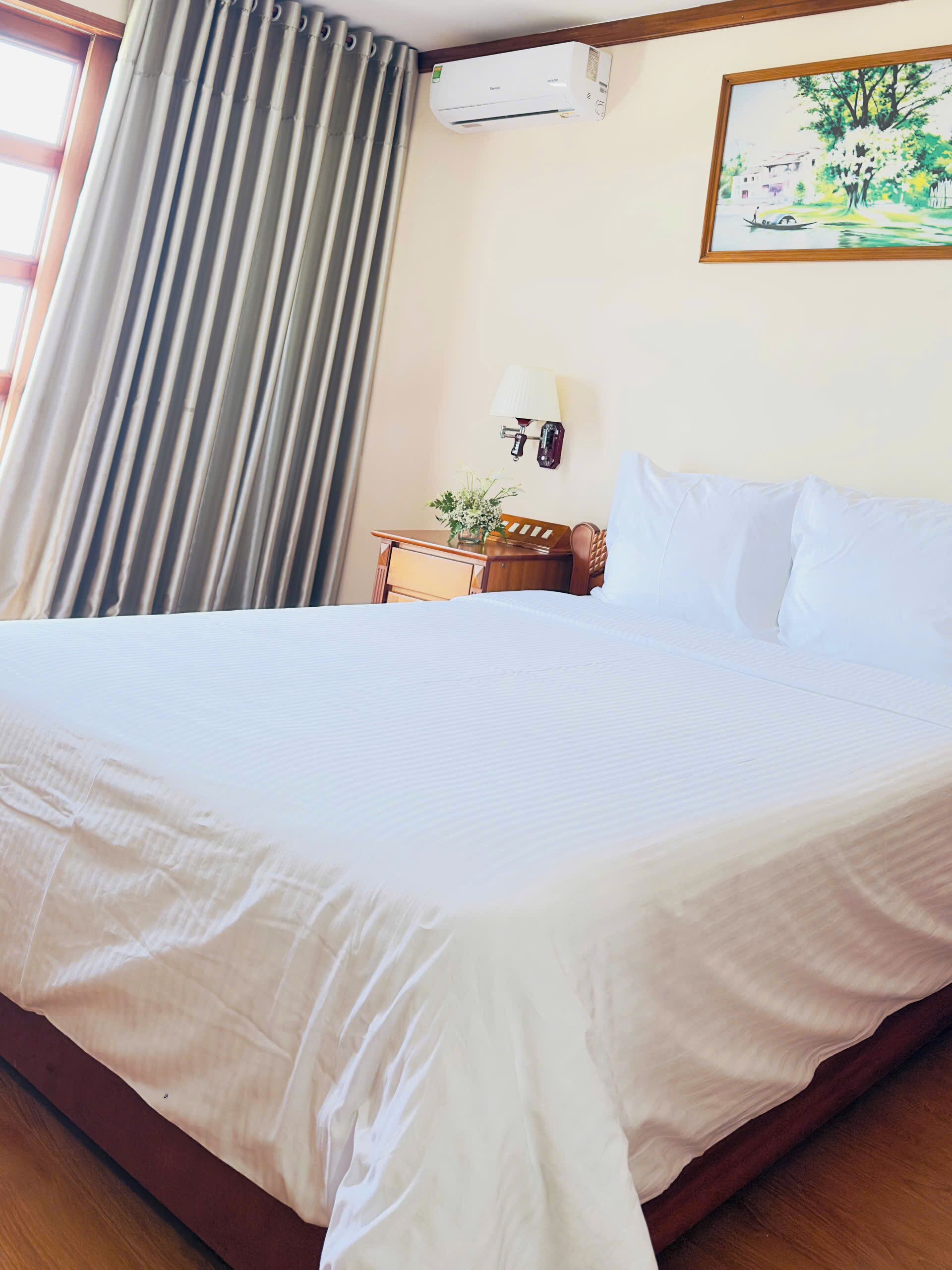 A hotel room with a large bed, a nightstand with a lamp, a wall-mounted air conditioner, and a painting on the wall.