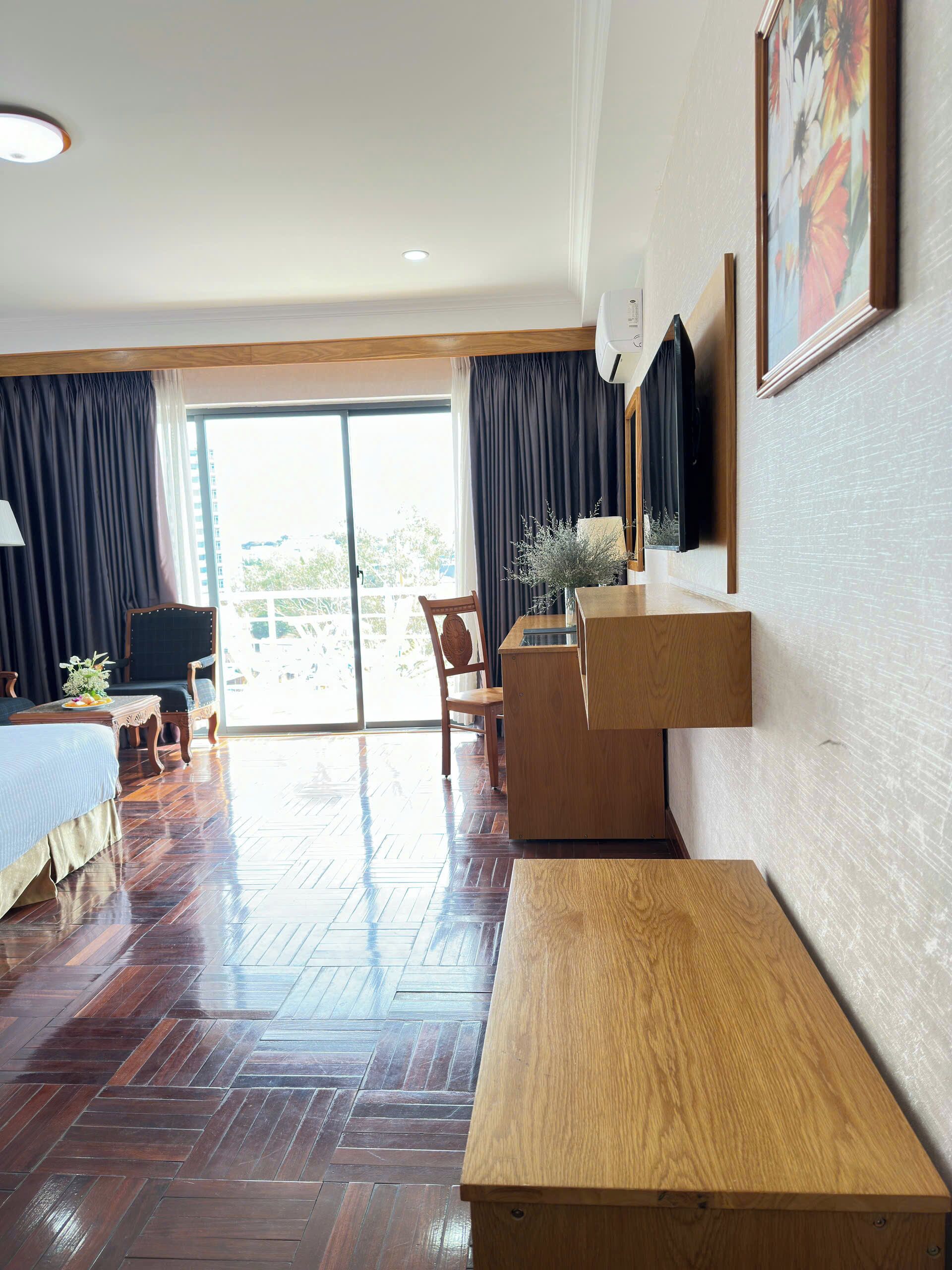 Une chambre d’hôtel avec un plancher en bois, un lit, un bureau et une chaise. Il y a une fenêtre avec des rideaux et une vue sur l’extérieur.
