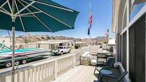 Una terraza con mobiliario exterior, un espacio cubierto con sombrilla, una bandera y vista a una carretera con vehículos y un parque de trailers al fondo.