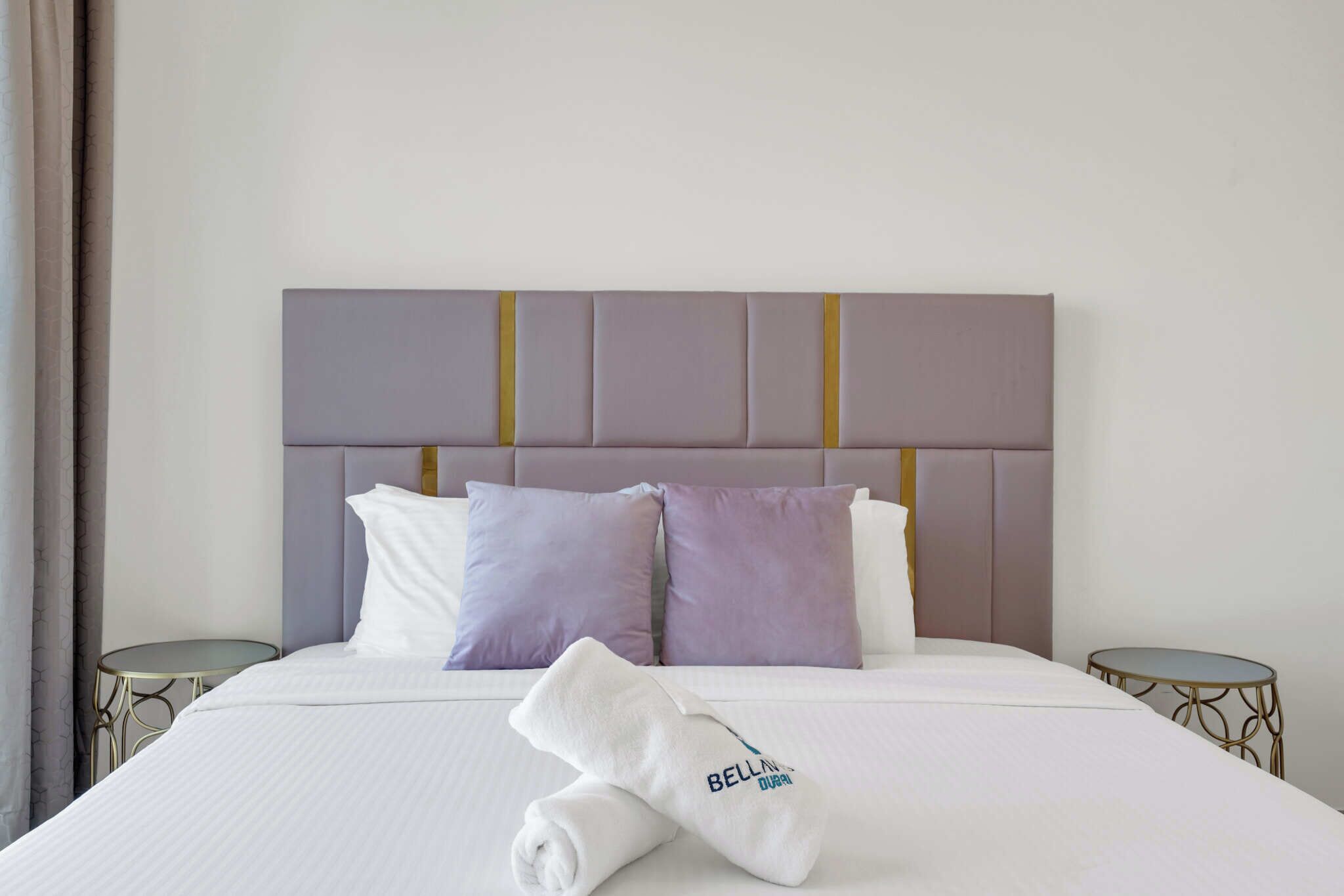 A neatly made bed with a white and purple pillow set, a folded towel with "BELLINGDURAN" on it, and two small round tables on either side.