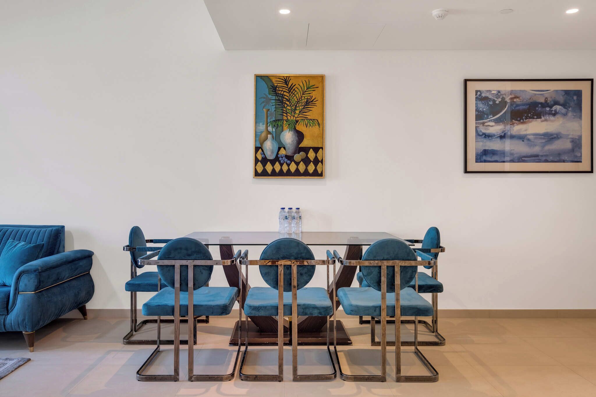 A modern living room with a blue sofa, a dining table with four blue chairs, and two paintings on the wall.