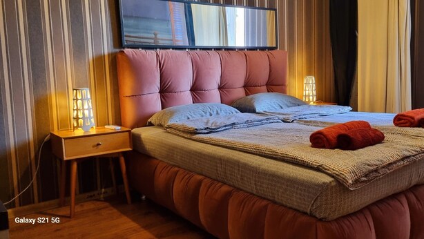 Moderni makuuhuone, jossa on napitettulla päällistyksellä varustettu sängynpääty, sängyn yläpuolelle asennettu litteänäyttöinen televisio ja kaksi yöpöytävalaisinta.