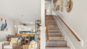 Una cocina moderna con armarios blancos, un comedor con mobiliario de mimbre y una escalera con barandilla de madera.