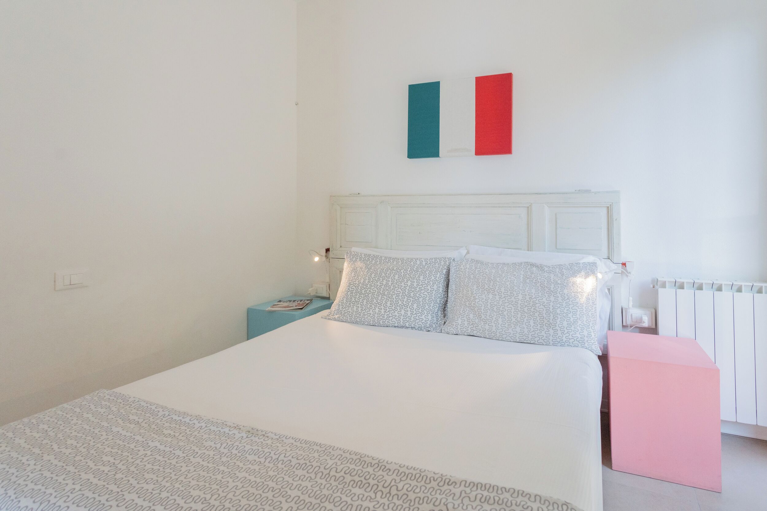 Une chambre avec un lit blanc, une table de chevet, une petite table d’appoint rose et un radiateur.
