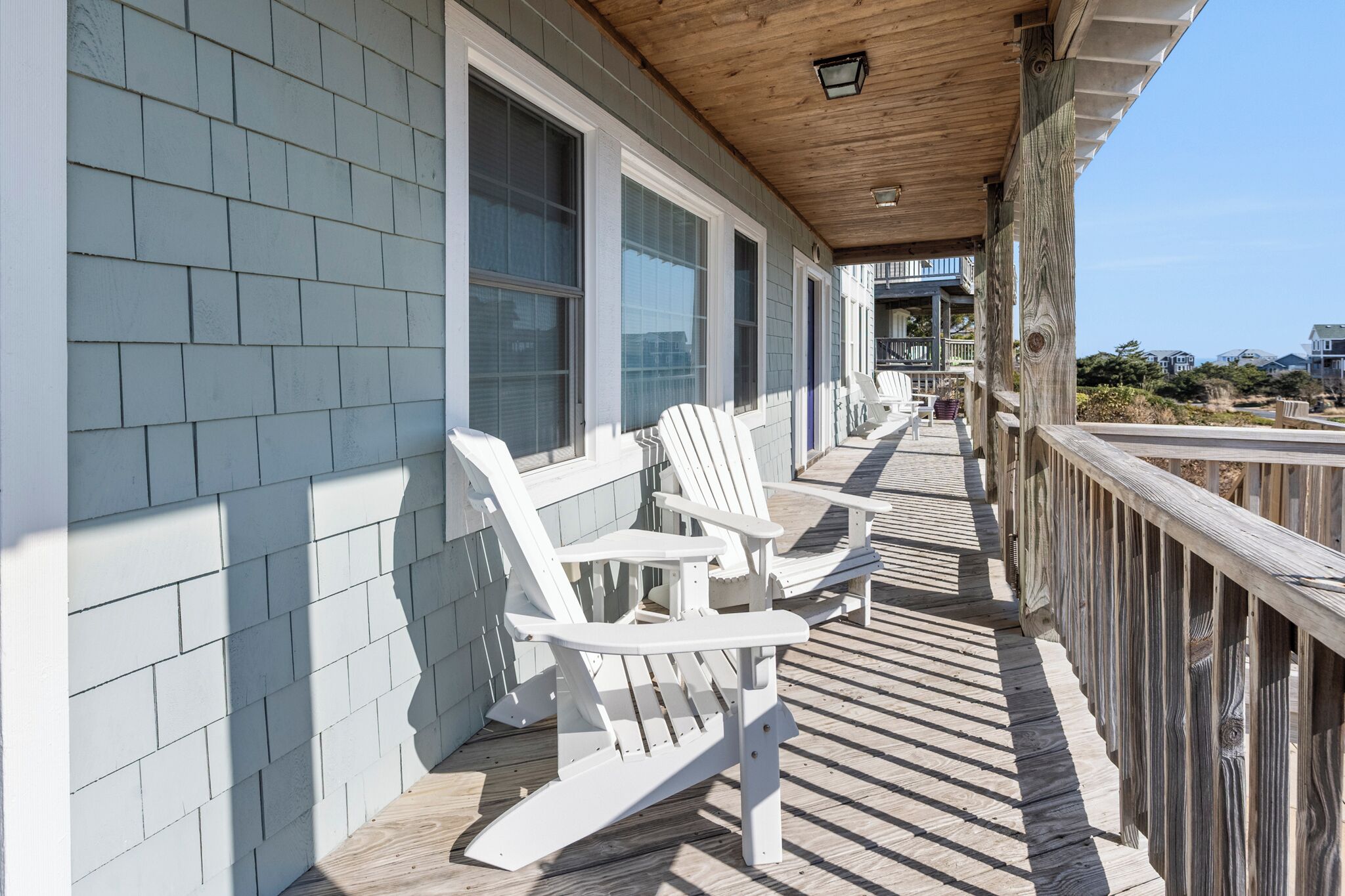Une terrasse en bois avec des fauteuils Adirondack blancs, une balustrade en bois et une vue sur l’océan.