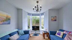 Een woonkamer met twee blauwe banken, een tv op een dressoir, een kroonluchter en een erker met een schilderij.