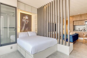 A modern bedroom with a large bed, a glass shower enclosure, and a kitchenette in the background.