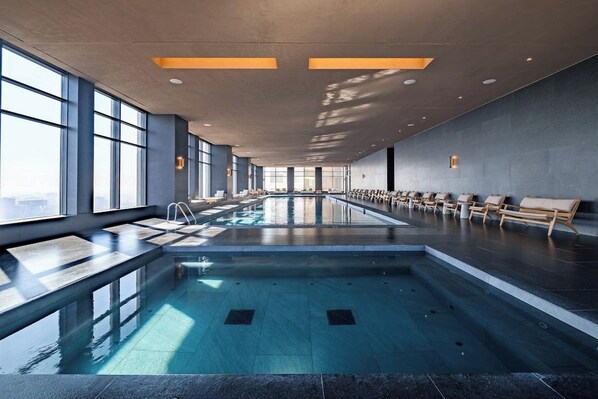 A large indoor swimming pool with a long bench seating area along one side and a sunlit ceiling.
