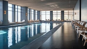 Een groot binnenzwembad met loungebedden en ligstoelen langs de randen, grote ramen die voor natuurlijk licht zorgen, en een modern architectonisch ontwerp.