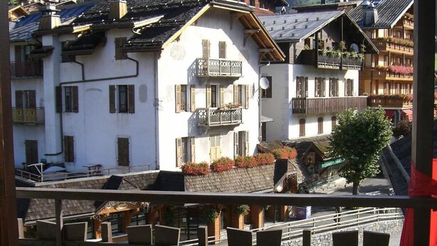 A balcony with a white chair and a small table, overlooking a village with traditional buildings and a mountainous backdrop. - 50m from resort center, 4th floor, balcony, 24m², Le Grand Bornand (Le Grand-Bornand)