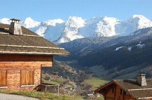 A wooden chalet with a sloped roof, a chimney, and a view of snow-capped mountains in the background.