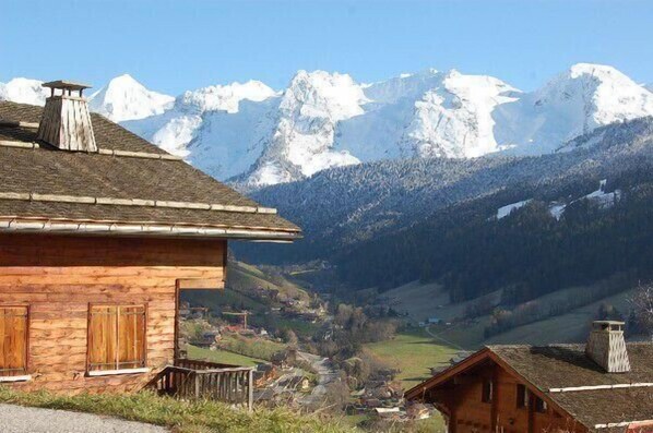 A wooden chalet with a sloped roof, a chimney, and a view of snow-capped mountains in the background.