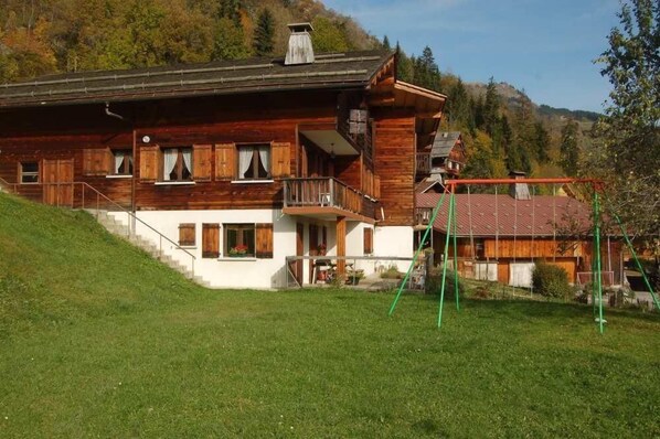 A wooden chalet with a balcony, a staircase leading to the entrance, and a red-roofed structure nearby.