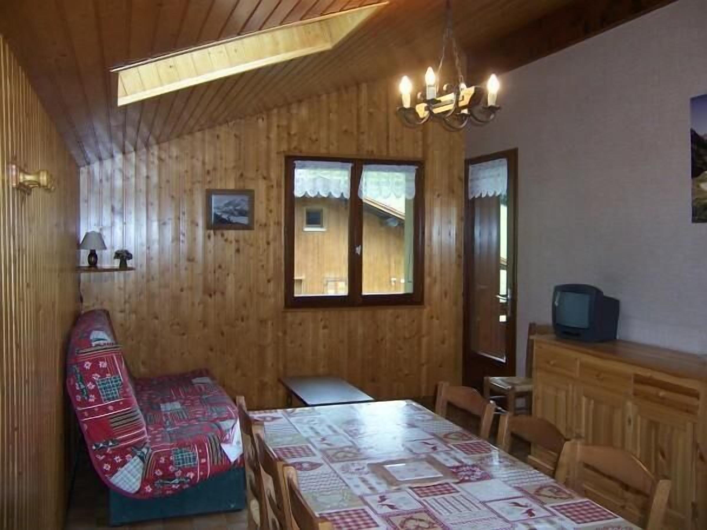 Interno di una baita in legno con tavolo, sedie, una panca, un lampadario e un televisore.