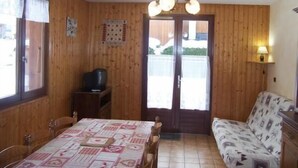 Ein gemütliches Zimmer mit Holzwänden, einem Esstisch mit gemustertem Tischläufer, einer Couch mit gemustertem Bezug, einer Deckenleuchte und einer Tür, die nach draußen führt.
