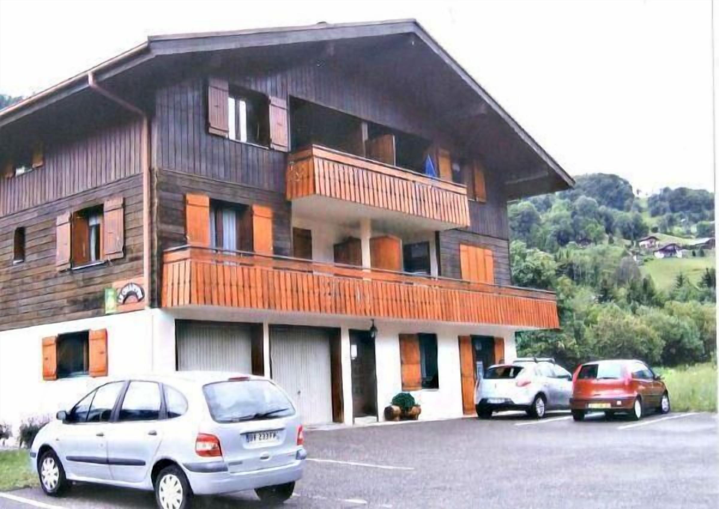 A two-story building with a wooden facade, balconies, and a parking area with cars.