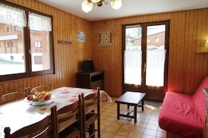 Ein gemütliches Zimmer mit Holzwänden, einem Esstisch mit Obstschale, einer Couch und einem Fernseher auf einer Holzkommode.