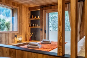 A wooden cabin room with a bed, towels, shelves with decorative items, and a view of trees through the window.