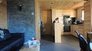 L’intérieur d’un chalet moderne présente un mur en pierre, un plafond en bois, un canapé en cuir noir, une table à manger et un coin cuisine.