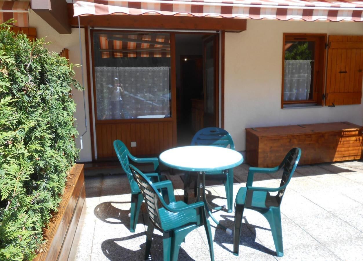An outdoor patio with a round table and four chairs, a wooden bench, and a small window with a curtain.