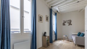 Une pièce comprenant un canapé, une petite table et un radiateur. Il y a deux fenêtres avec des rideaux bleus, et un mur orné de deux tableaux encadrés.