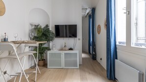 Un salon moderne avec un téléviseur, un coin repas comprenant des chaises et une table, une plante et une grande fenêtre avec des rideaux.