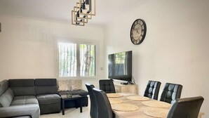 En moderne stue med en hjørnesofa, et spisebord med stole, et fjernsyn og et ur på væggen.