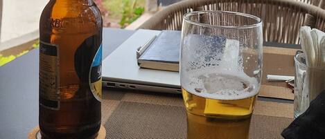 A table with a bottle and a glass of beer, a laptop, and a wicker chair.