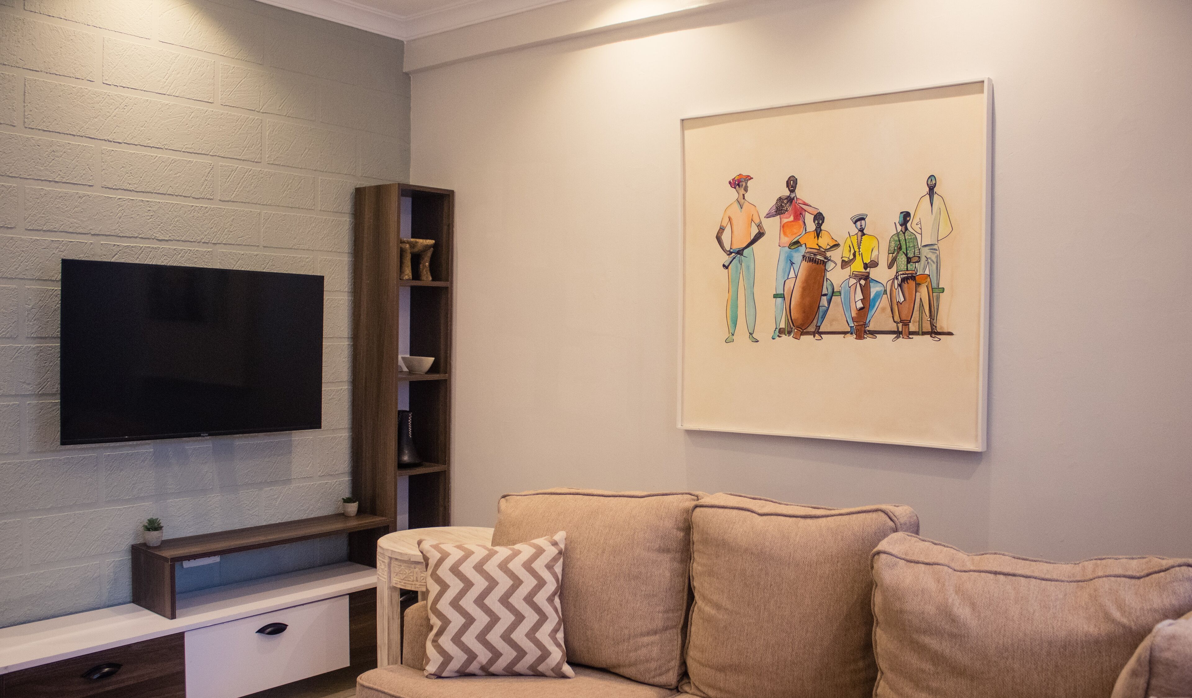 A living room with a flat-screen TV mounted on a textured wall, a wooden entertainment unit below, and a beige sofa with patterned cushions.