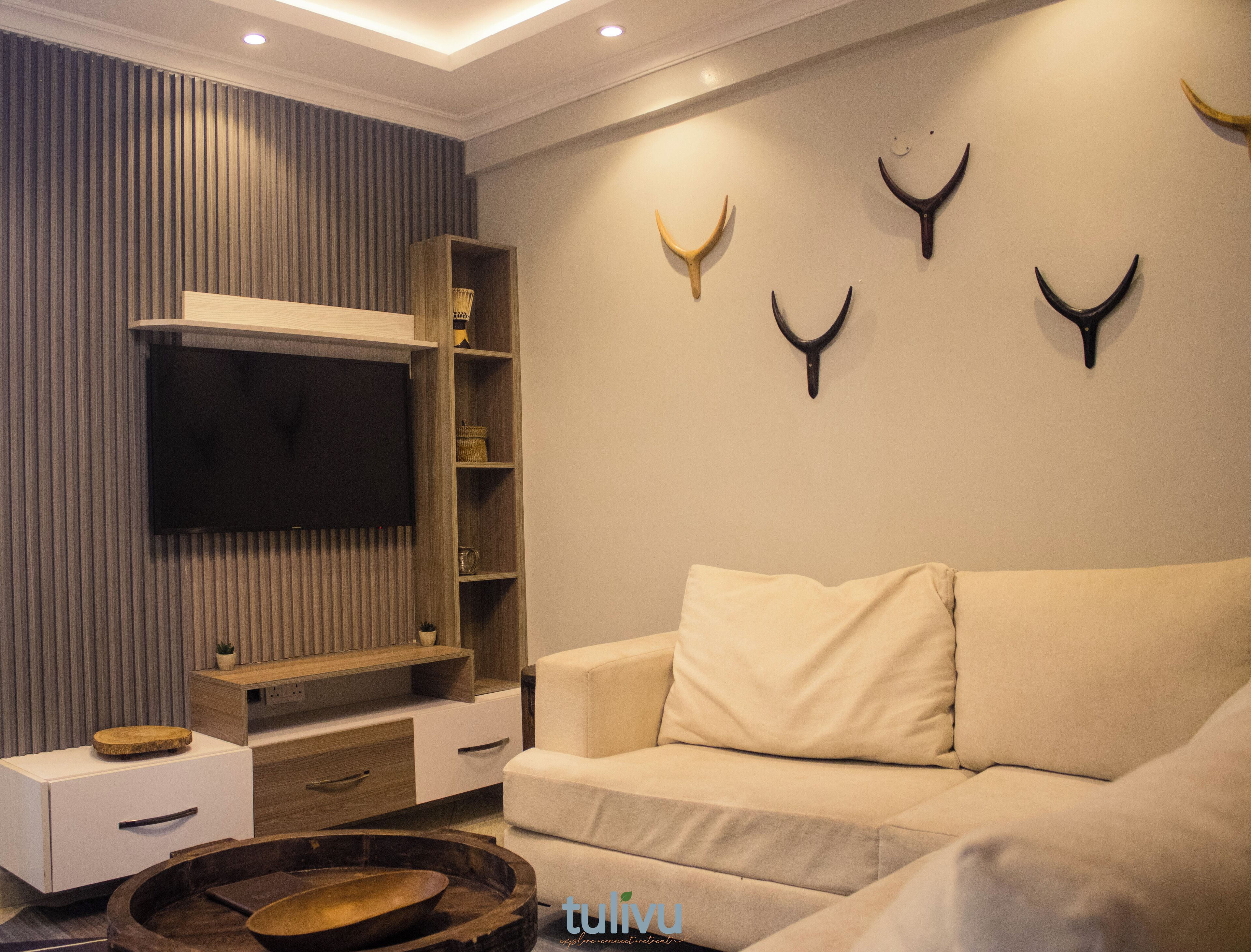 A modern living room with a flat-screen TV, wooden shelving, a beige sofa, and decorative antler wall art.