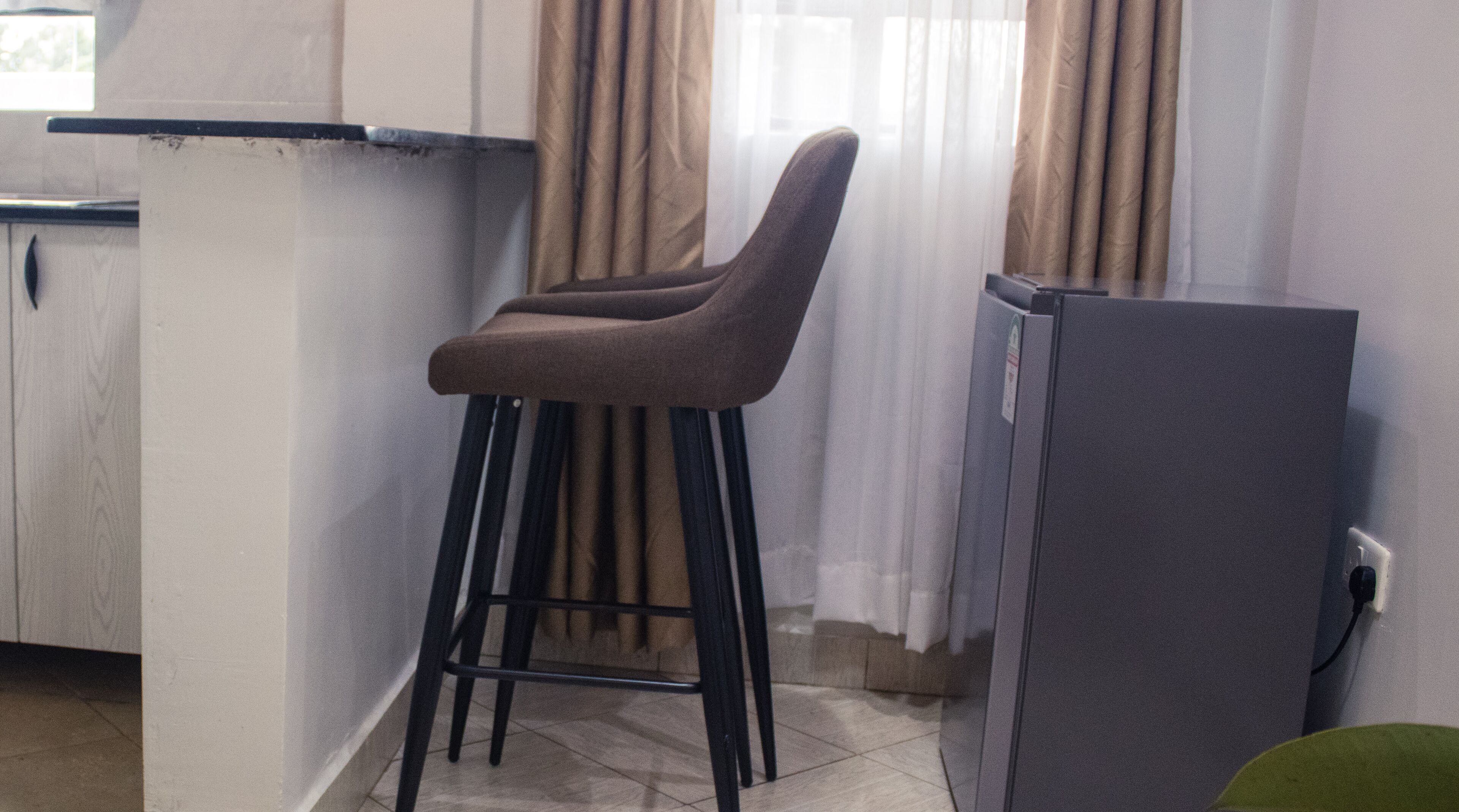 A kitchen area with a bar stool, a refrigerator, and a window with curtains.