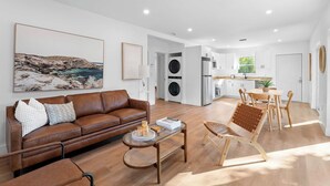 A modern living room with a brown leather sofa, a glass coffee table, and a dining area with wooden chairs. A large painting of a coastal landscape is on the wall. - Spacious, Bright Condo-Victoria Park Near Beach (Fort Lauderdale)