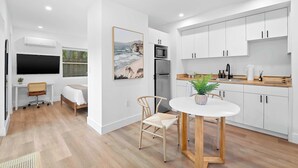 A modern kitchen with white cabinets, a dining table, and a large window overlooking a beach scene. - Modern Clean Studio Apartment Near Beach (Fort Lauderdale)