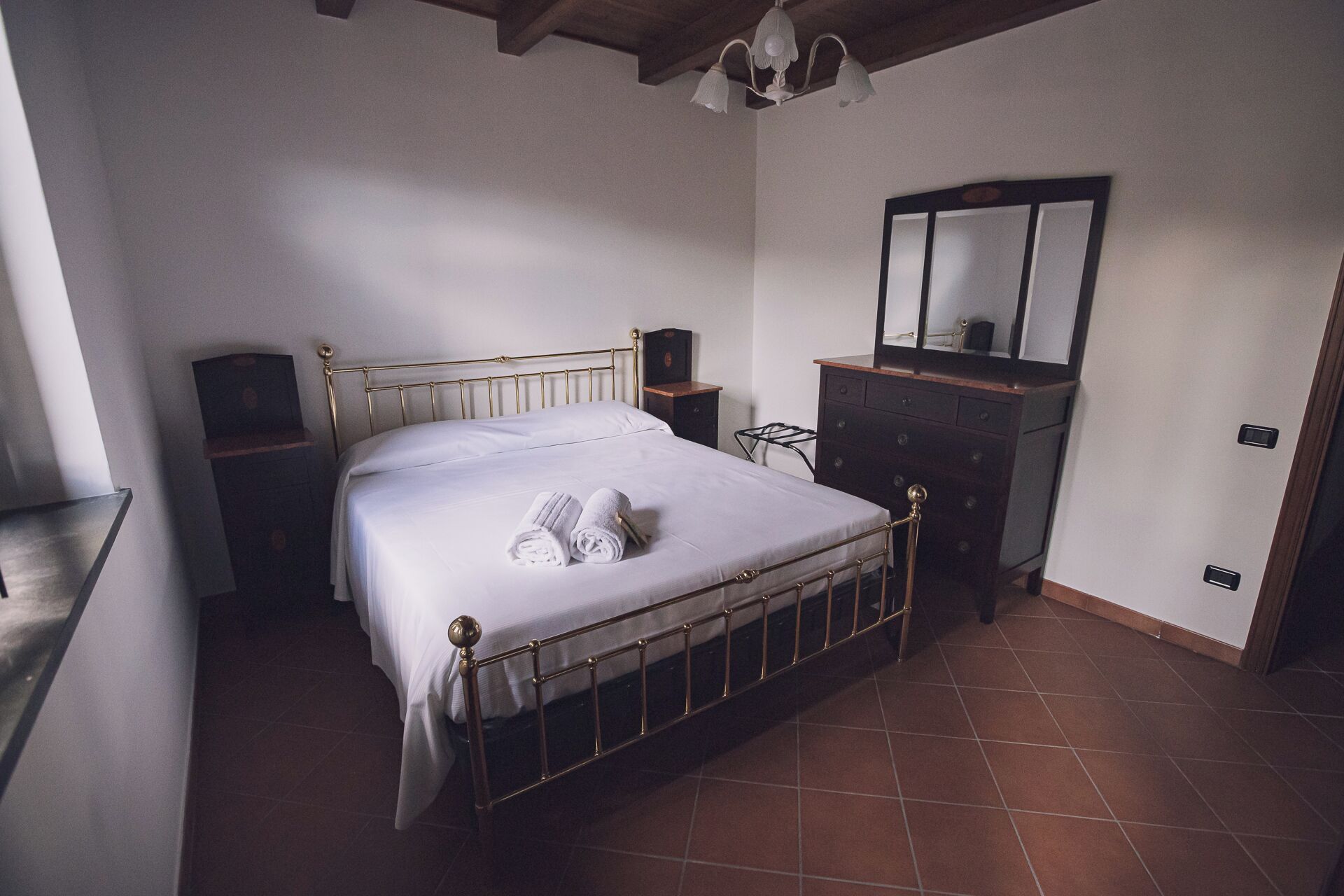 Quarto com cama de estrutura metálica, cómoda de madeira, espelho e uma pequena mesa de cabeceira.