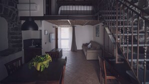 A spacious loft with a high ceiling, exposed wooden beams, a dining area with a table and chairs, a sofa, and a staircase with a metal railing. - LA GELSOMINA (Mascali)