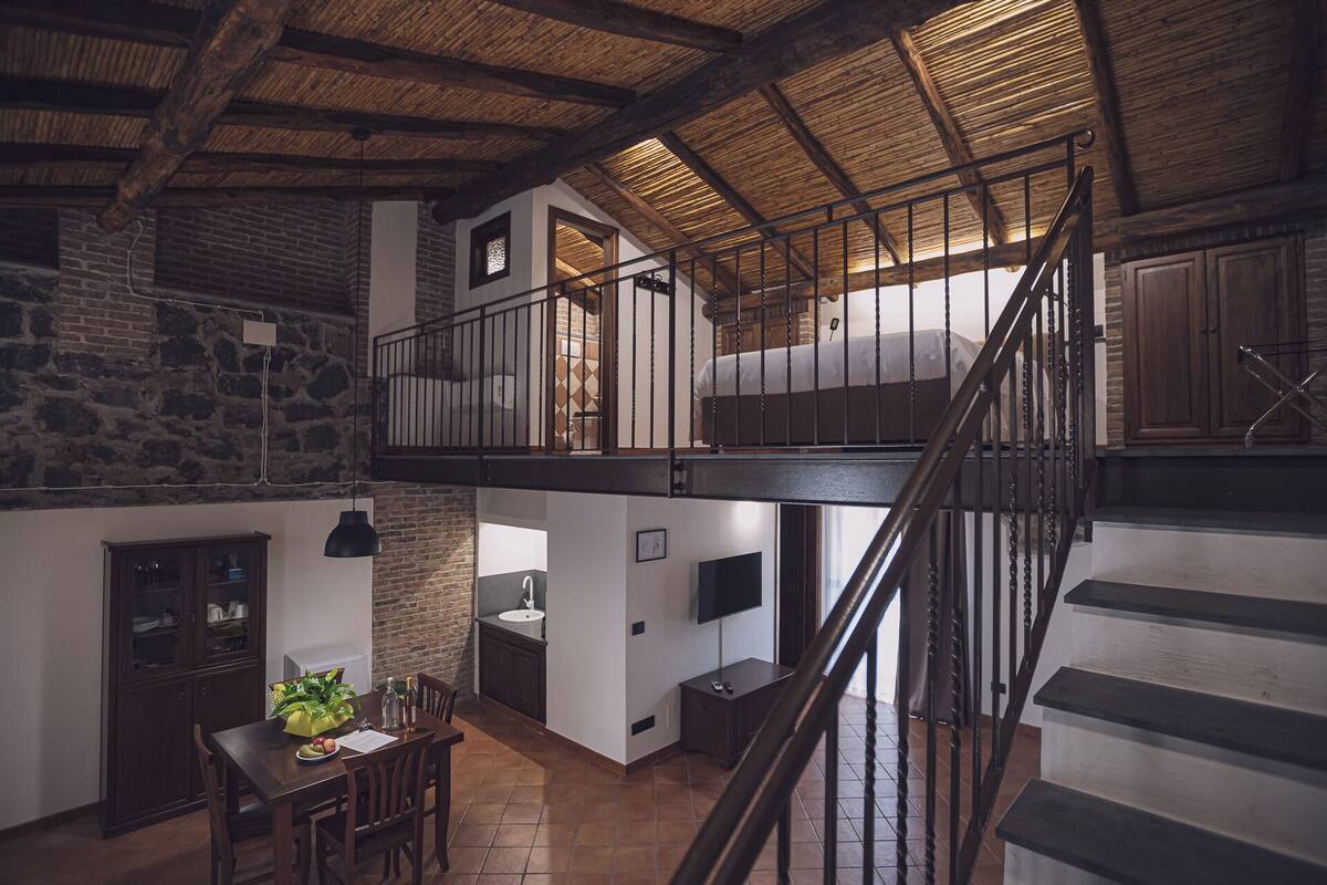 Un loft espacioso con vigas de madera a la vista, cama de altillo, zona de comedor con mesa y sillas, y cocina con pileta y encimera.