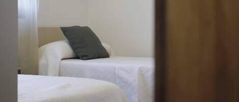 Een kamer met twee bedden, elk met witte lakens en een grijs kussen. De kamer heeft een houten plafond en een effen witte muur.