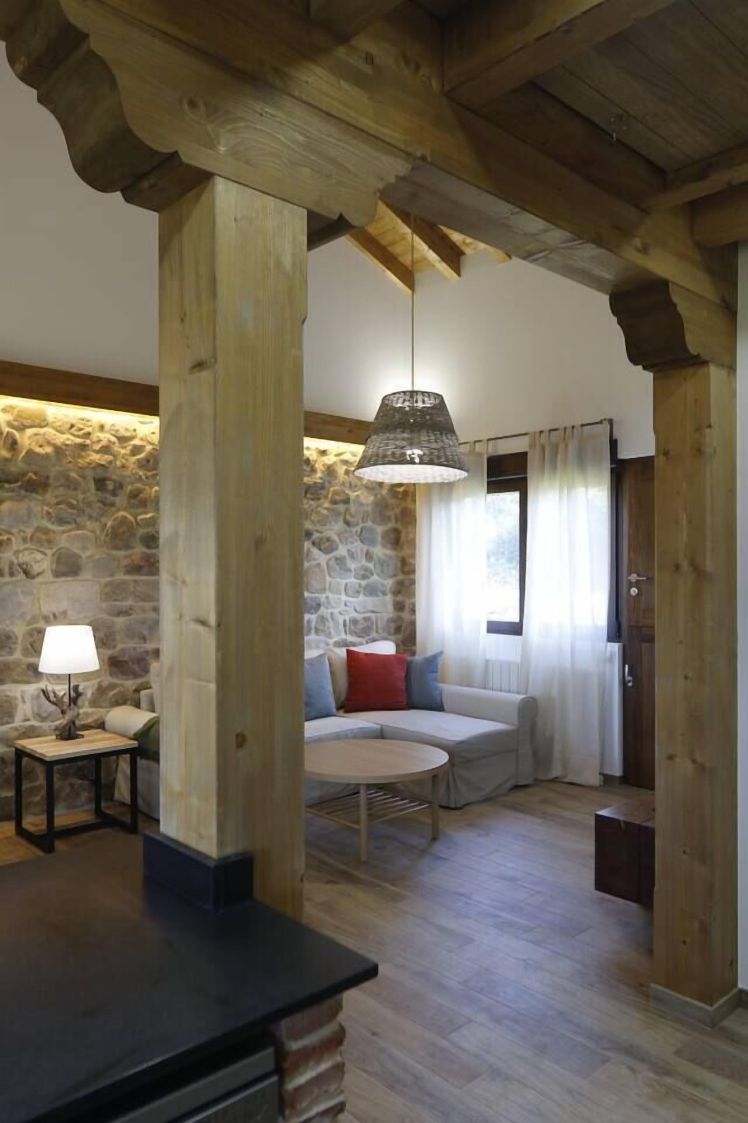 A cozy living room with a stone wall, wooden beams, a sofa with cushions, a table, and a hanging lamp.